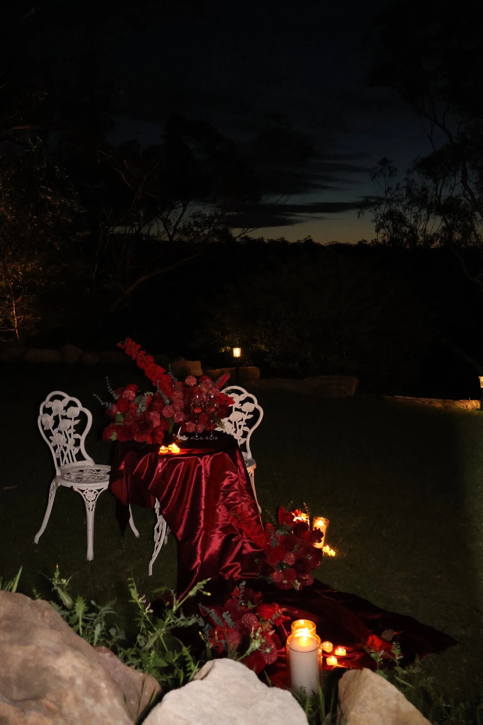 A romantic outdoor setup at night with a small round table draped in a red satin cloth, surrounded by two white vintage garden chairs. The table is decorated with red flowers, candles, and wine glasses, creating an intimate atmosphere. Candles are also placed on the ground around the table, and the sky is dark with some clouds and silhouettes of trees in the background.