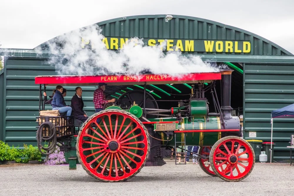 A vintage steam-powered tractor with red wheels and a black and green body on display outdoors. Three children are seated on the tractor, and steam is billowing from its chimney. A building with a sign reading "Pearn Steam World" is in the background.