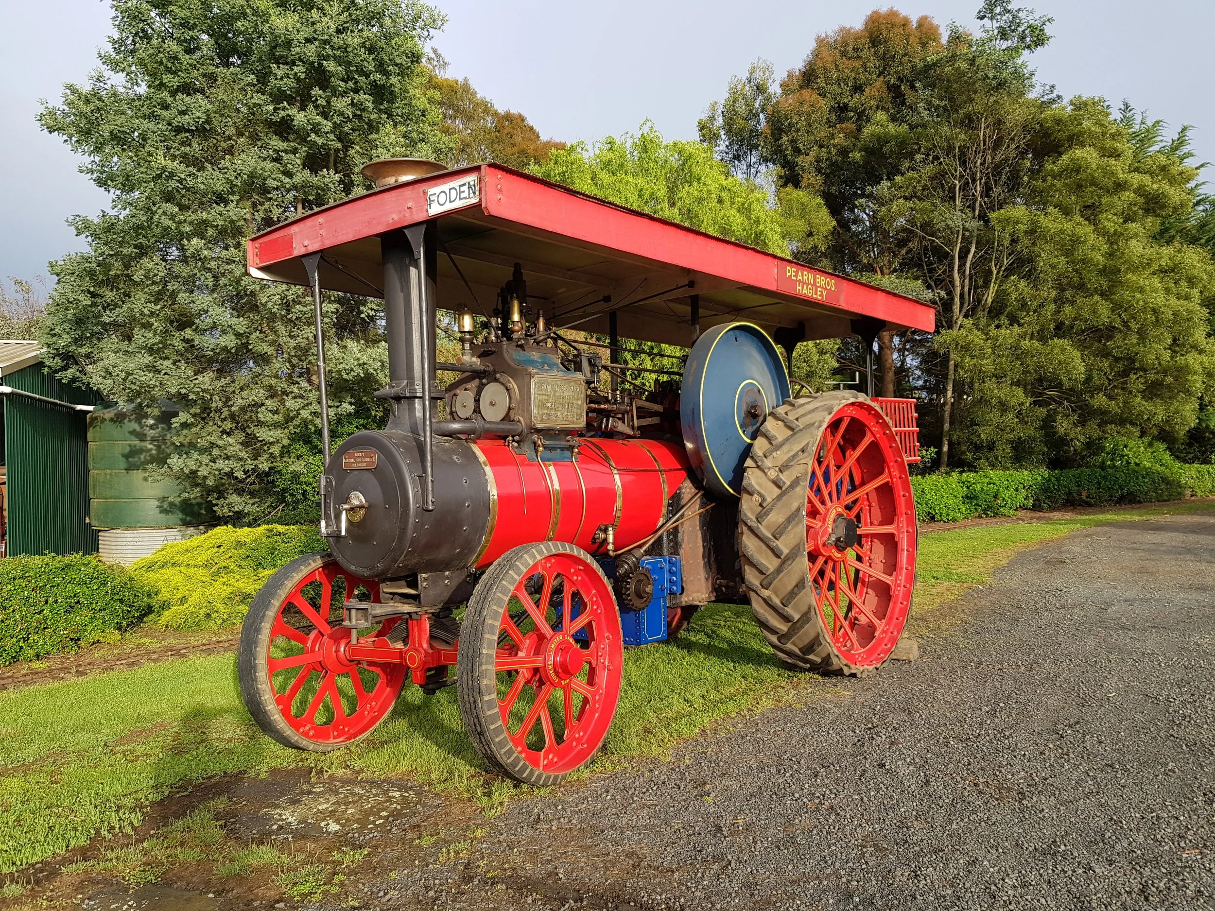 An old vintage steam tractor with red wheels, black and red body, and a blue pulley, parked on a patch of grass next to a gravel road, with trees and bushes in the background.