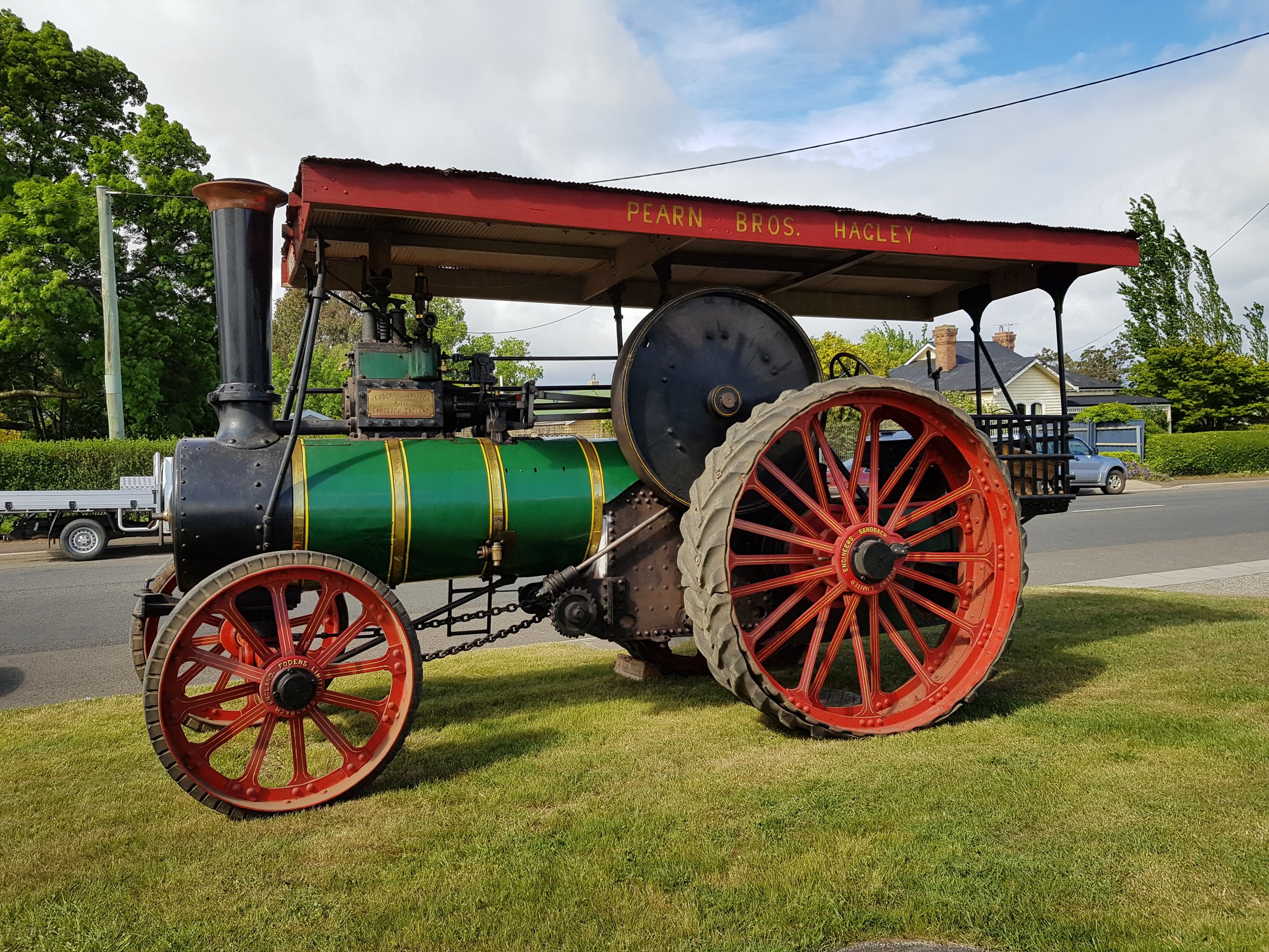 Vintage steam-powered tractor with large red wheels on a grassy area, with trees, a road, and houses in the background.