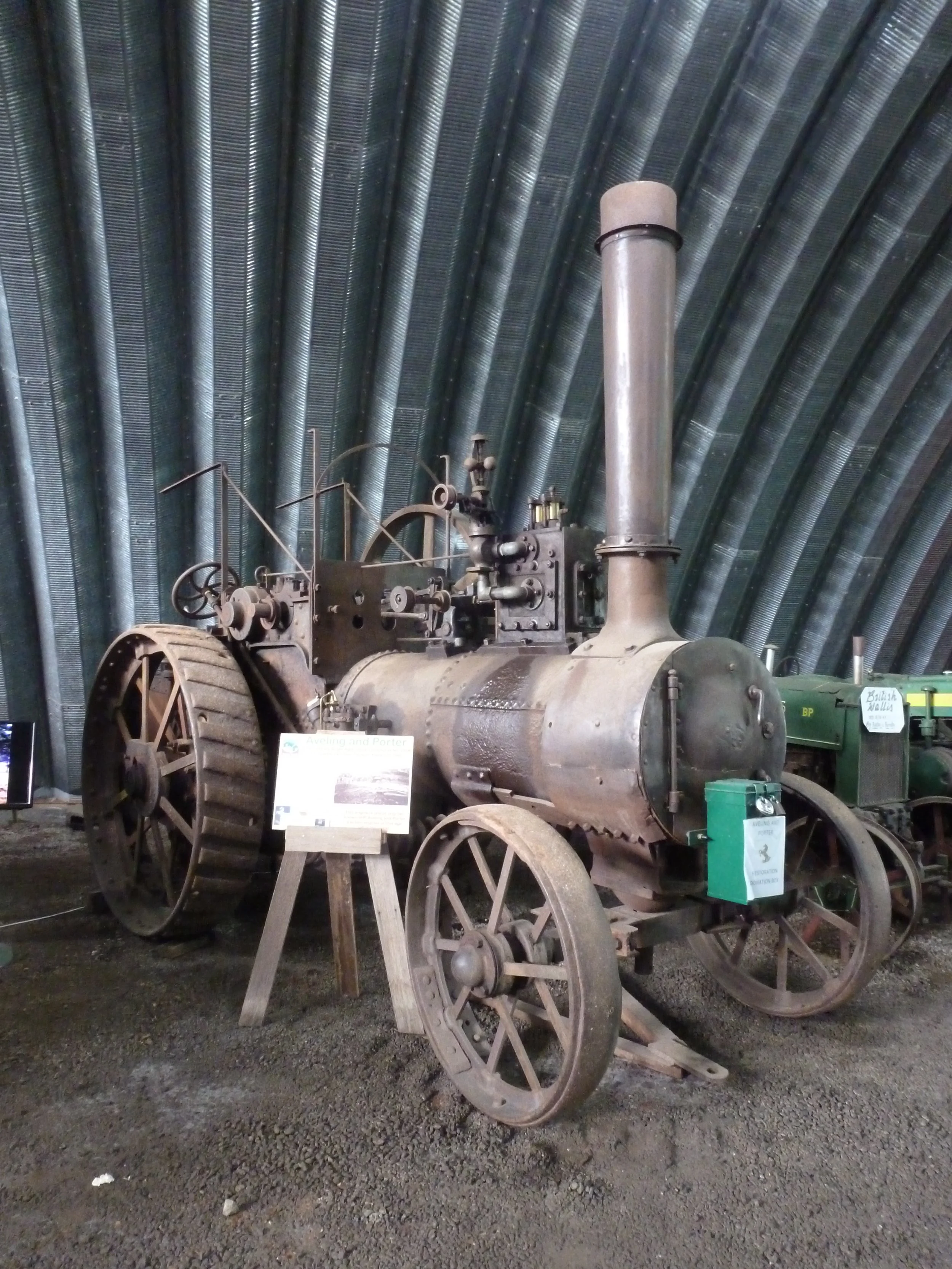 An old, large steam-powered tractor with big metal wheels and a tall chimney, displayed indoors with gravel floor and curved metal ceiling.