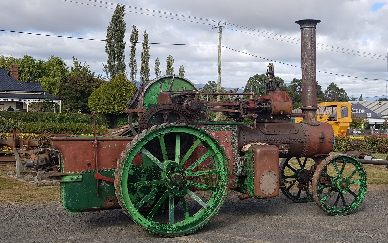 Old, rusty steam-powered traction engine with green wheels on a gravel surface, with trees, a house, and a cloudy sky in the background.