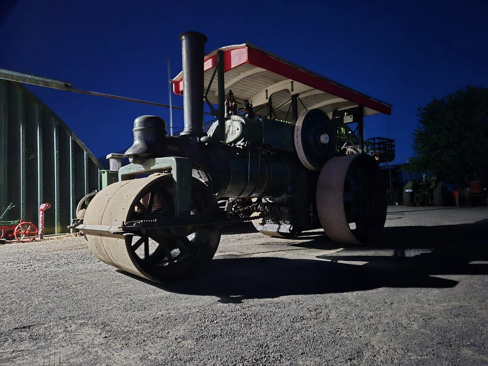 Vintage steamroller with large front and rear wheels parked on a gravel surface at night, with a blue sky and some trees and buildings in the background.