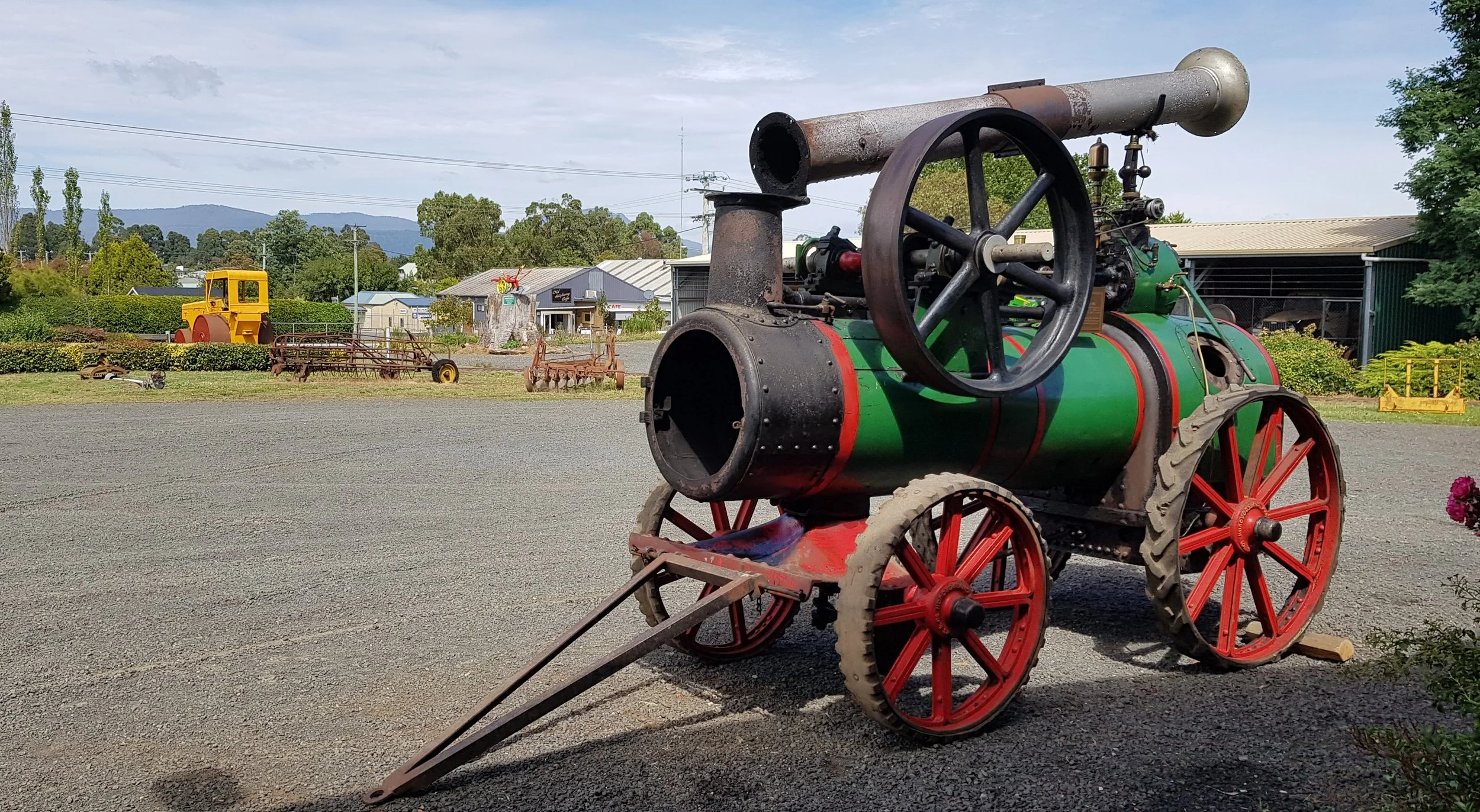 An old-fashioned steam-powered machine with large red wheels and a green body, situated outdoors on gravel, with farm equipment and buildings in the background, and a partly cloudy sky overhead.