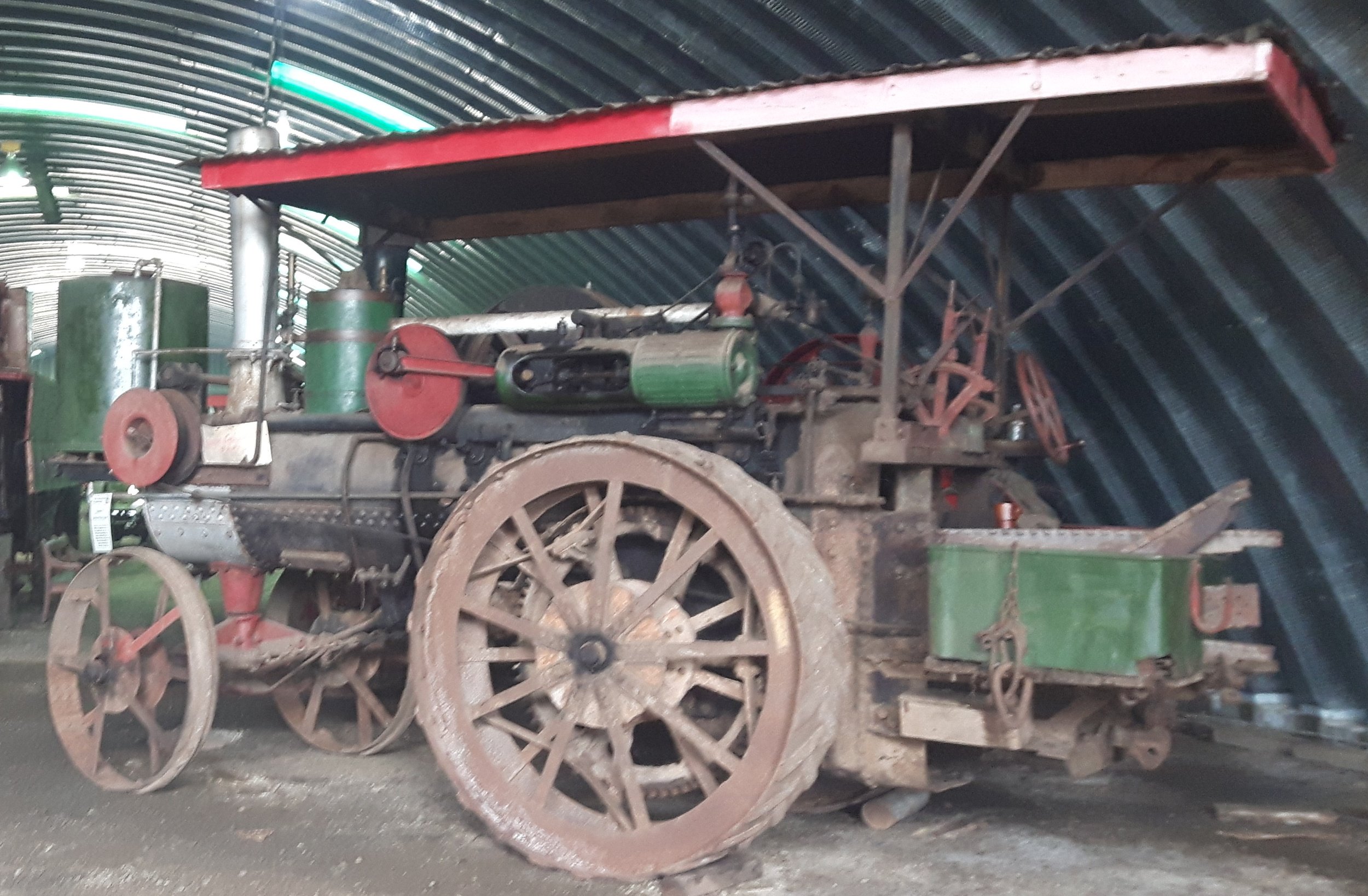 An old, rustic steam-powered tractor with large spoked wheels, painted in green and red, displayed in a museum with a curved, metal ceiling.