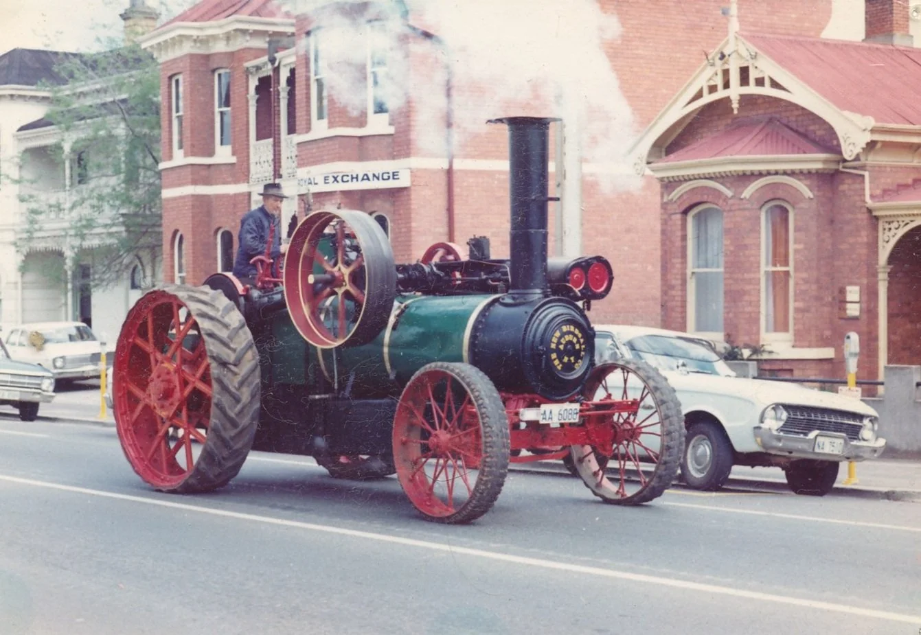 A vintage steam-powered tractor on a city street, with a man wearing a hat operating it, and brick and Victorian-style buildings in the background.