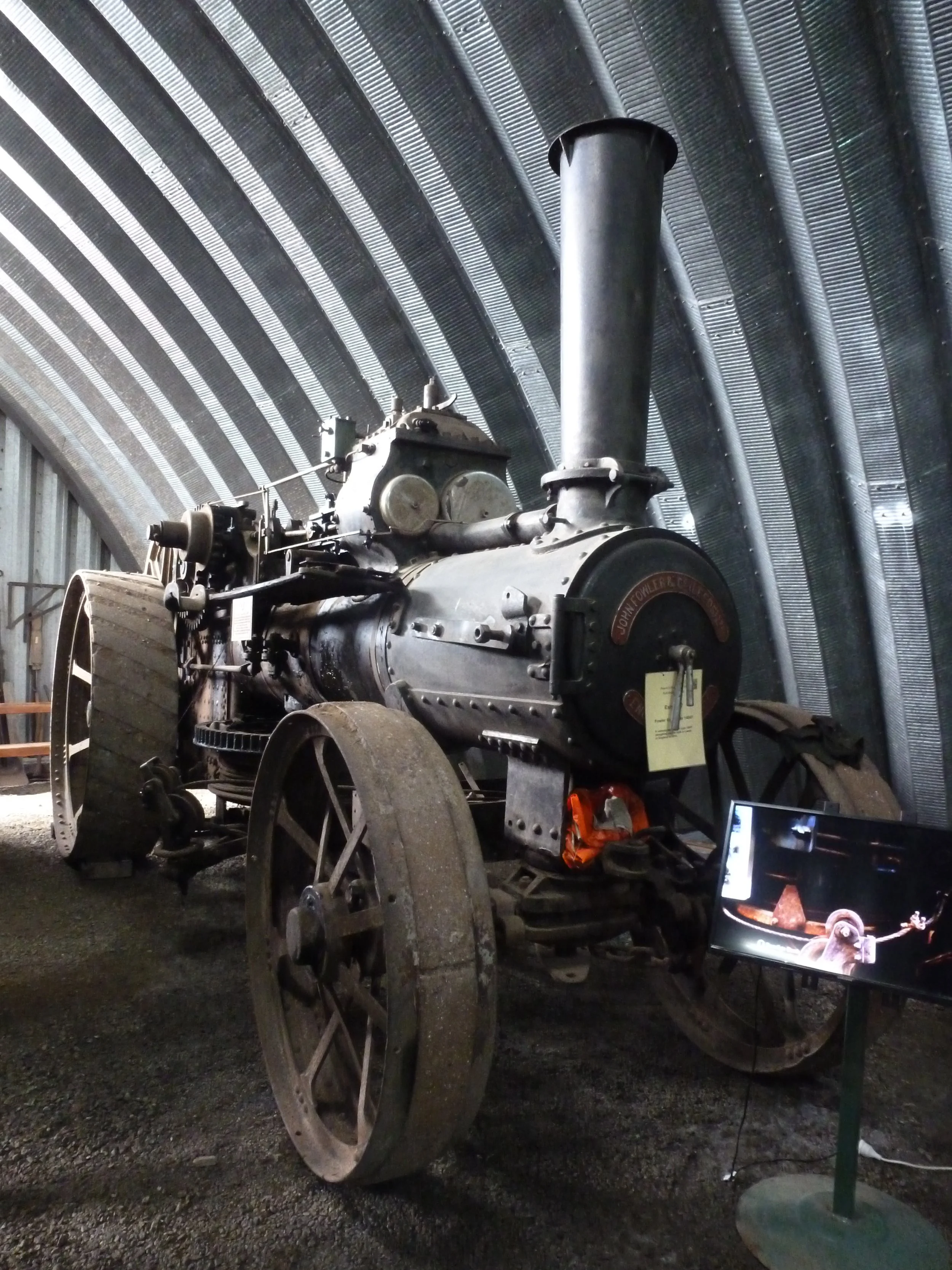 An old steam tractor with large wheels and a tall chimney, displayed in a museum with a curved metal ceiling and an informational screen beside it.