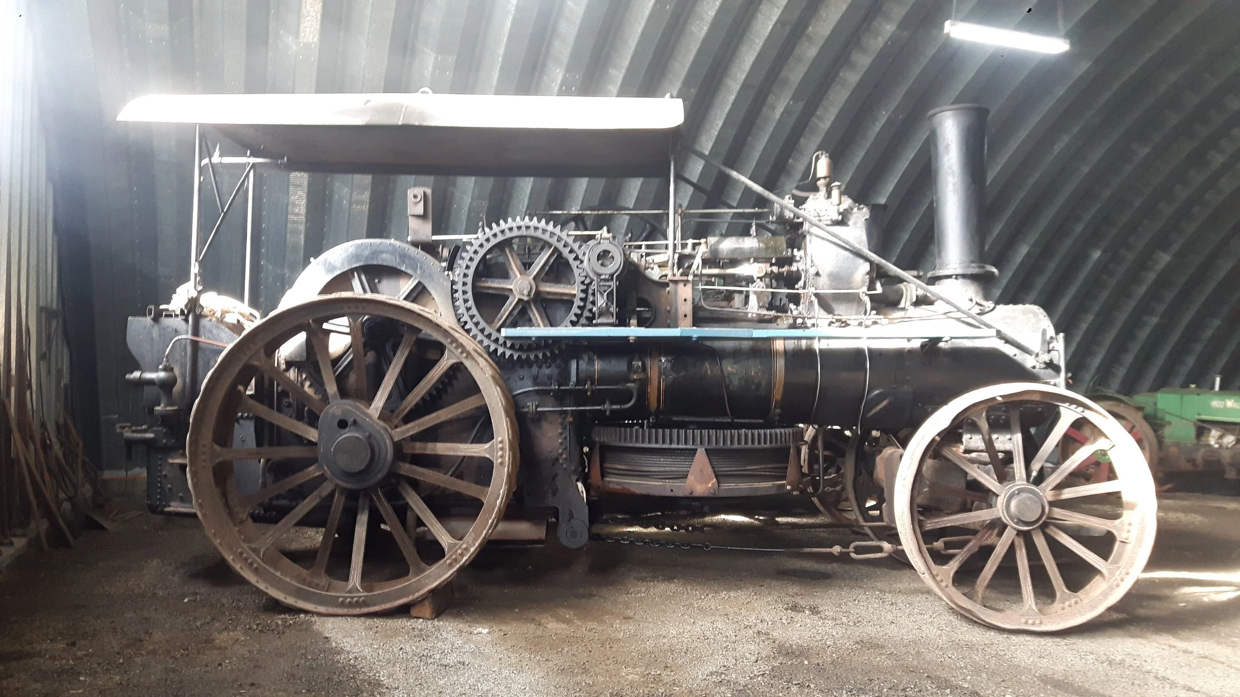 A vintage steam-powered tractor with large spoked wheels, exposed gears, and a chimney, displayed indoors against a metal wall.