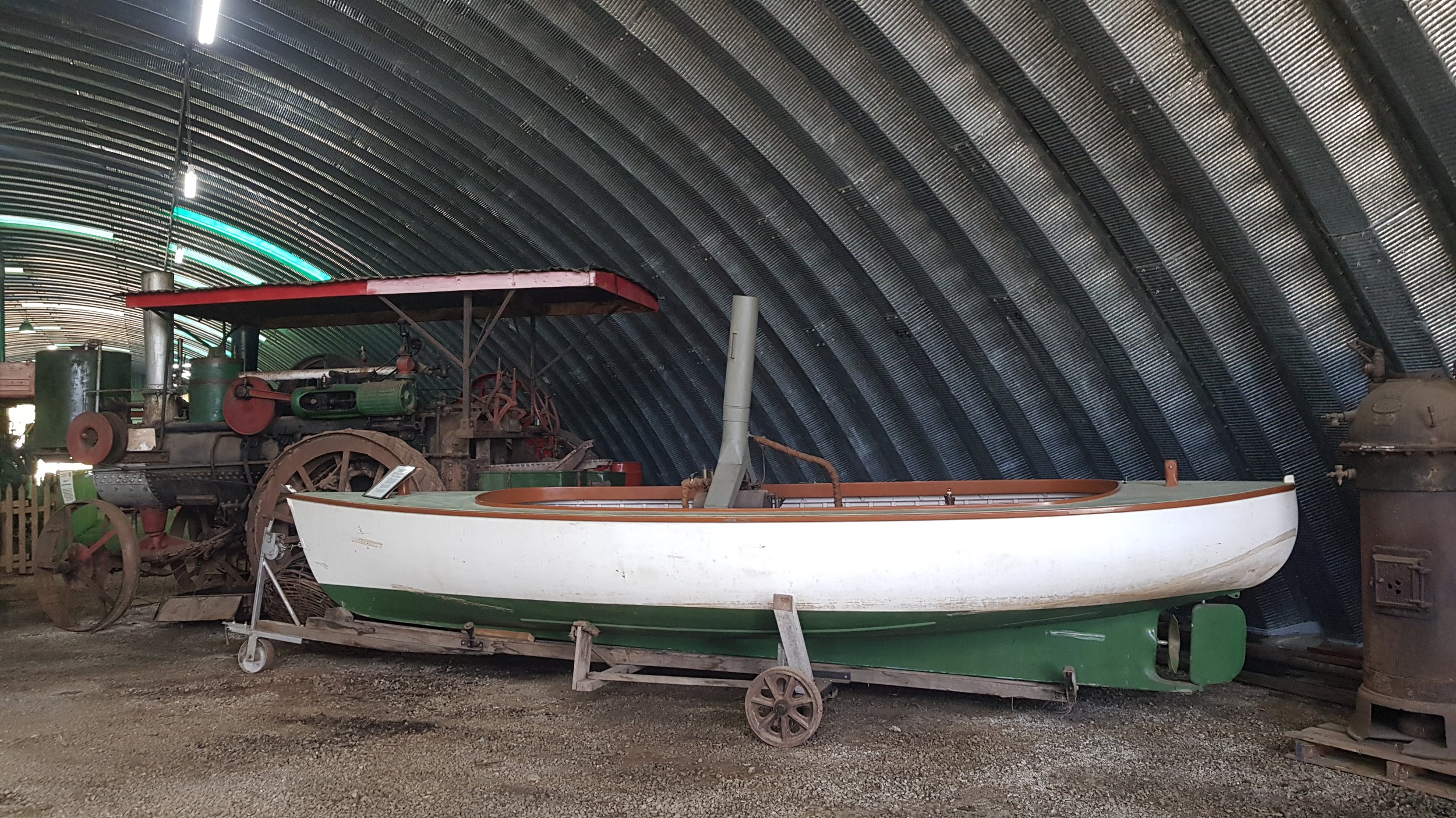 A vintage tractor and a small boat on display inside a curved metal building, with a gravel floor and some old equipment in the background.
