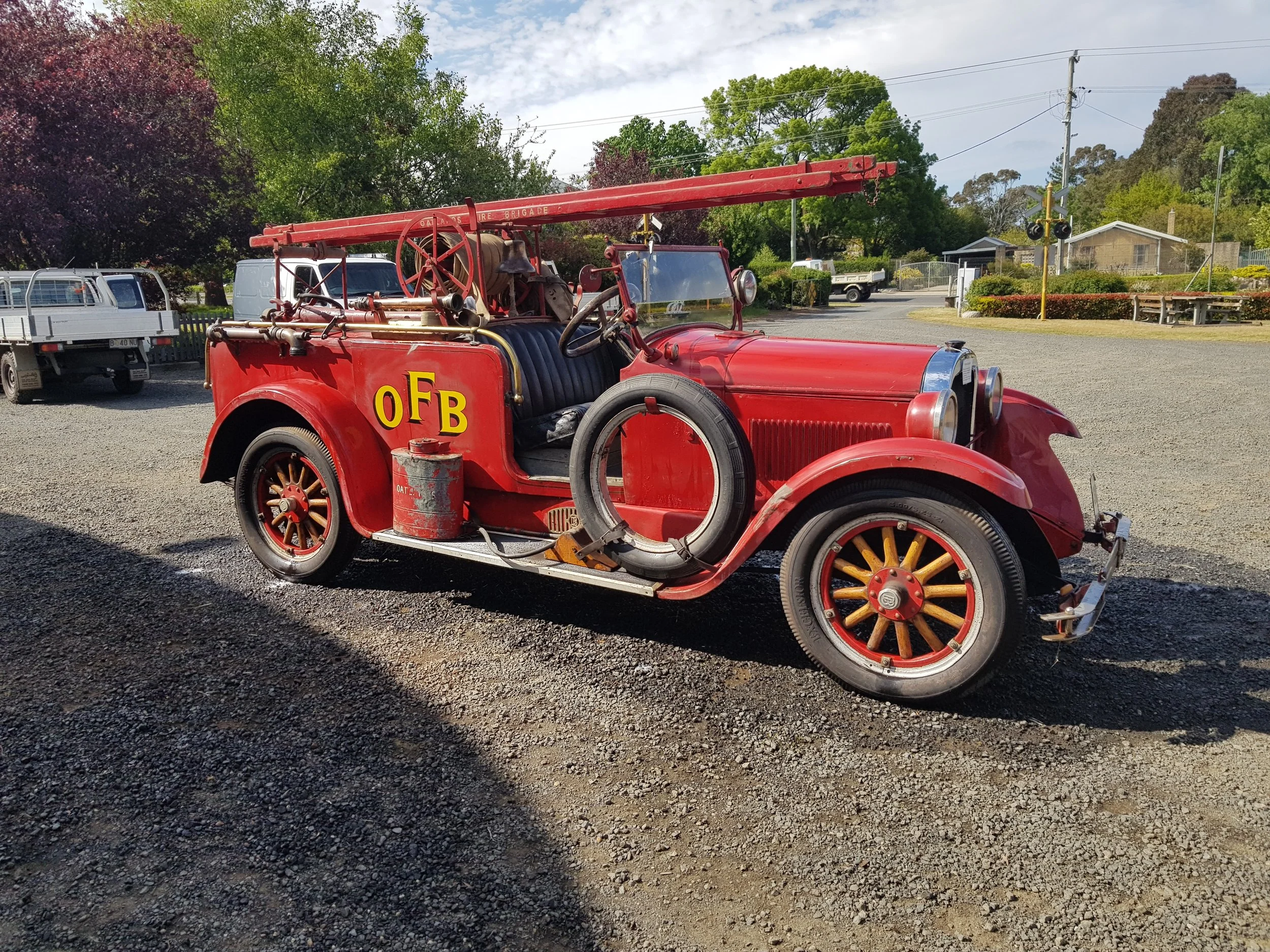 Vintage red fire truck with OFB letters, old wooden wheels, and firefighting equipment parked outdoors on gravel, surrounded by trees and other vehicles.