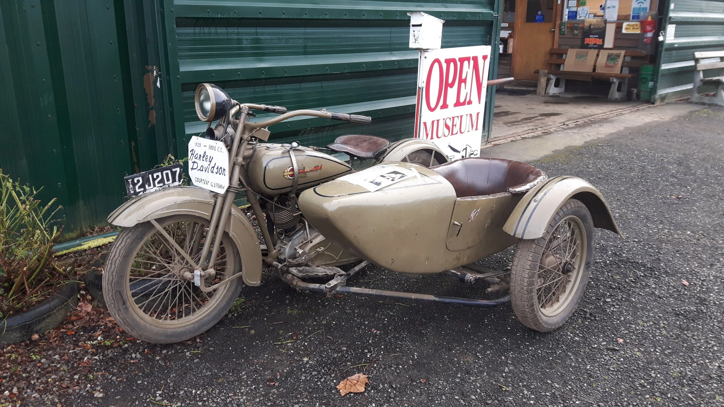 A vintage Harley Davidson motorcycle with a sidecar parked outside a museum. There is an 'Open Museum' sign nearby and a green building in the background.