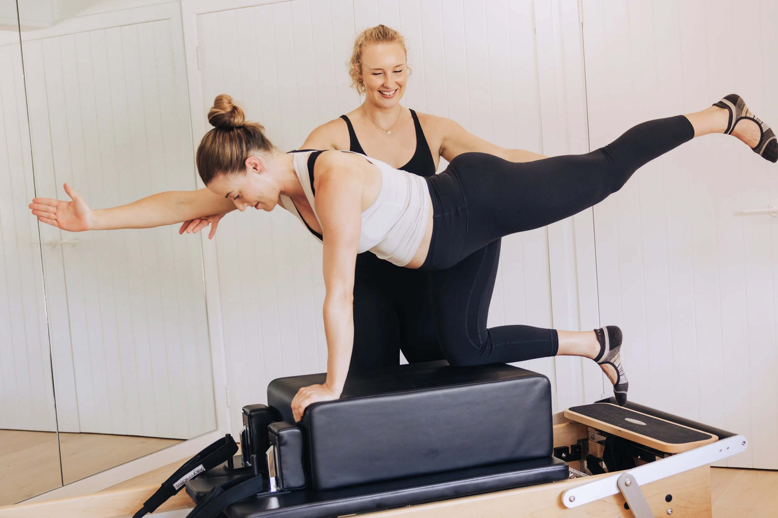 A woman in a white tank top and black leggings does a Pilates exercise on a reformer machine with assistance from an instructor in a black tank top in a fitness studio.