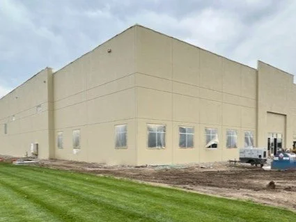 A large beige industrial building with multiple small windows, surrounded by grass and dirt, under a cloudy sky, with construction equipment in front.