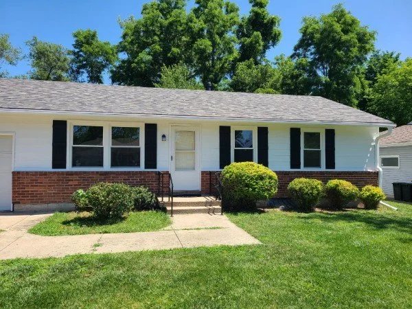 Front view of a single-story house with white exterior walls, black shutters, and a brick foundation. There are four bushes in front, a small concrete pathway leading to the front door, and tall trees in the background under a blue sky.
