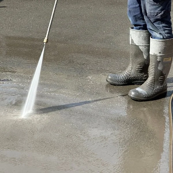 Person wearing rubber boots using a spray wand to apply a liquid onto a concrete surface.