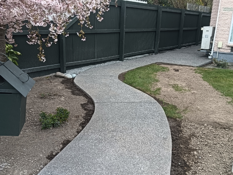 Curved concrete pathway in a backyard next to a black fence, with a garden bed, some grass, and a house with utility boxes.