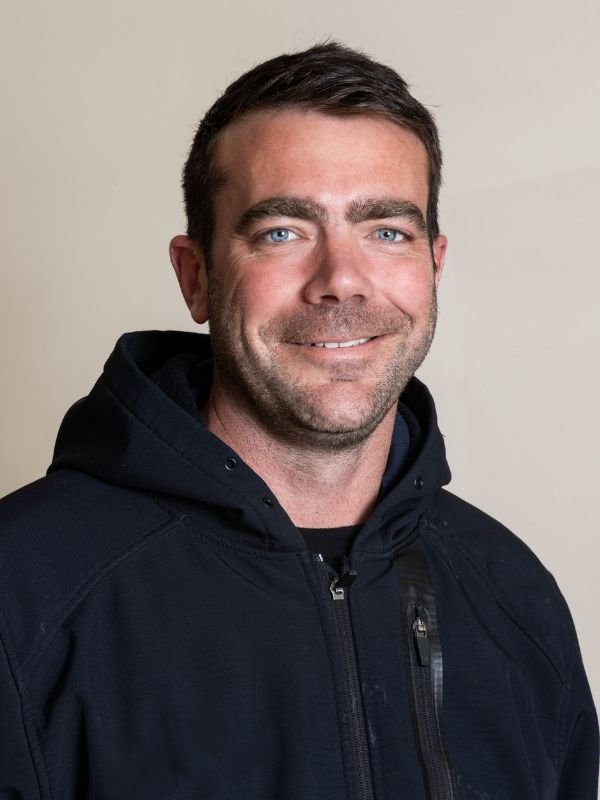 A man with short dark hair, blue eyes, and a beard, smiling while wearing a black zip-up hoodie against a light-colored background.