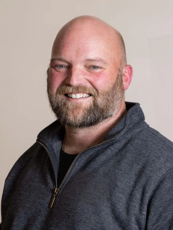 Headshot of smiling man with a beard and mustache, wearing a dark zip-up sweater, against a light background.