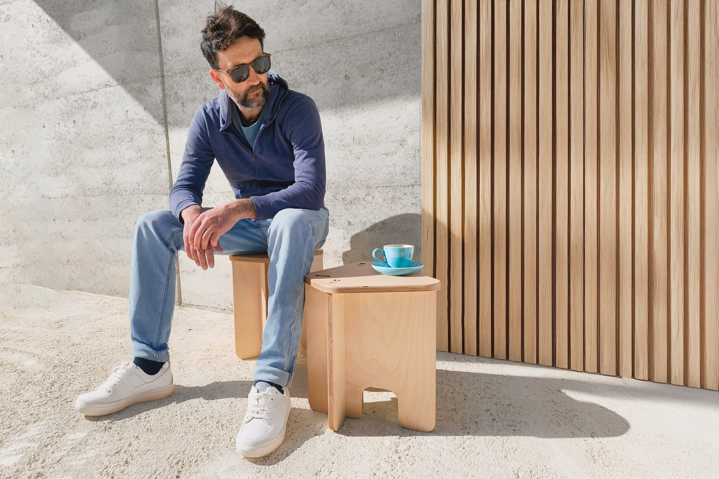 A man with dark hair, beard, wearing sunglasses, a navy blue hoodie, light jeans, and white sneakers sitting on a wooden stool outdoors. Beside him, there is a small wooden table with a blue cup and saucer. The background includes a concrete wall and a wooden slat fence, with sunlight casting shadows.