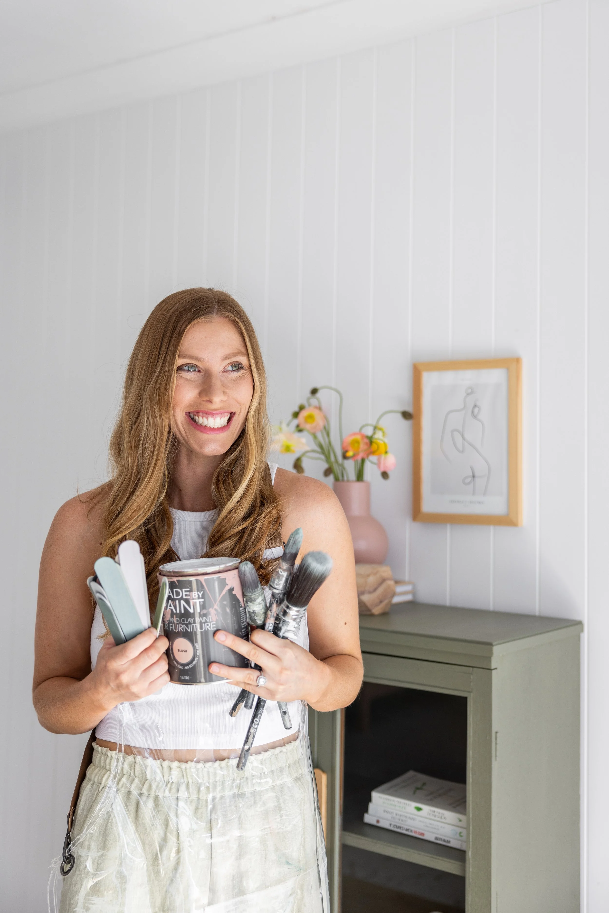 A woman smiling and holding a paint can and brushes in a room with white paneled walls, a light green cabinet, and a vase with pink and yellow flowers.
