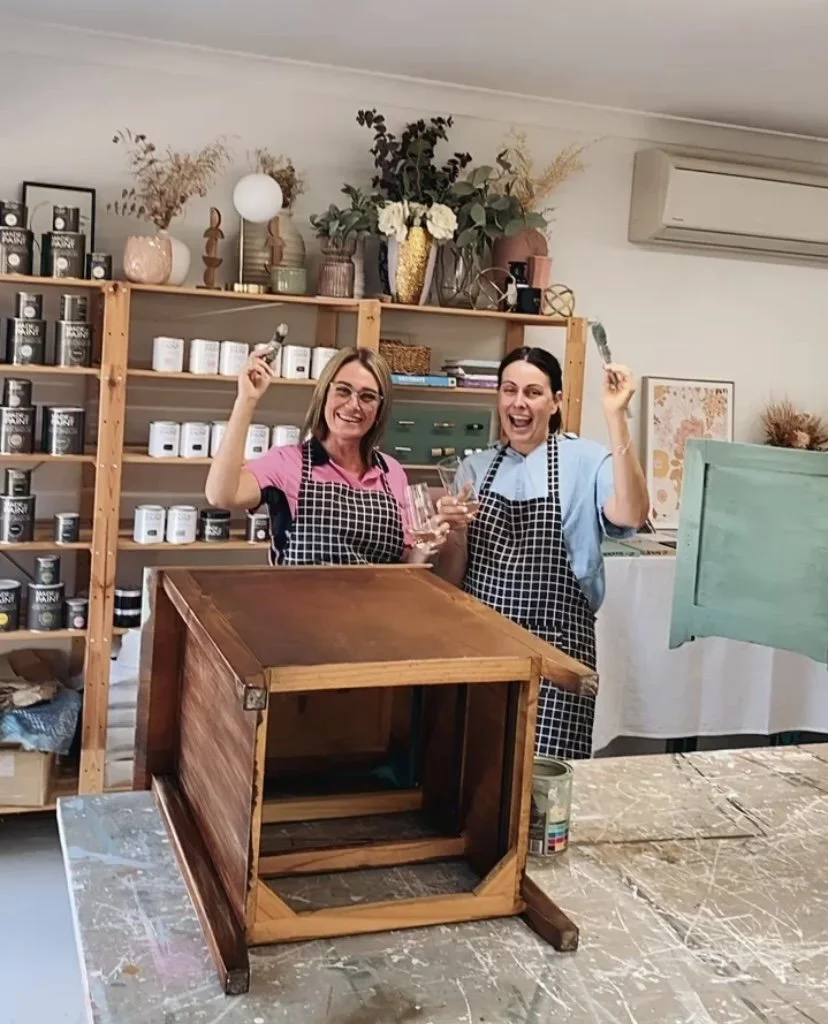 Two women wearing aprons are standing in a workshop, smiling and holding paintbrushes and glasses of wine. They are surrounded by shelves filled with jars and supplies, and there is a wooden piece of furniture on the table in front of them.
