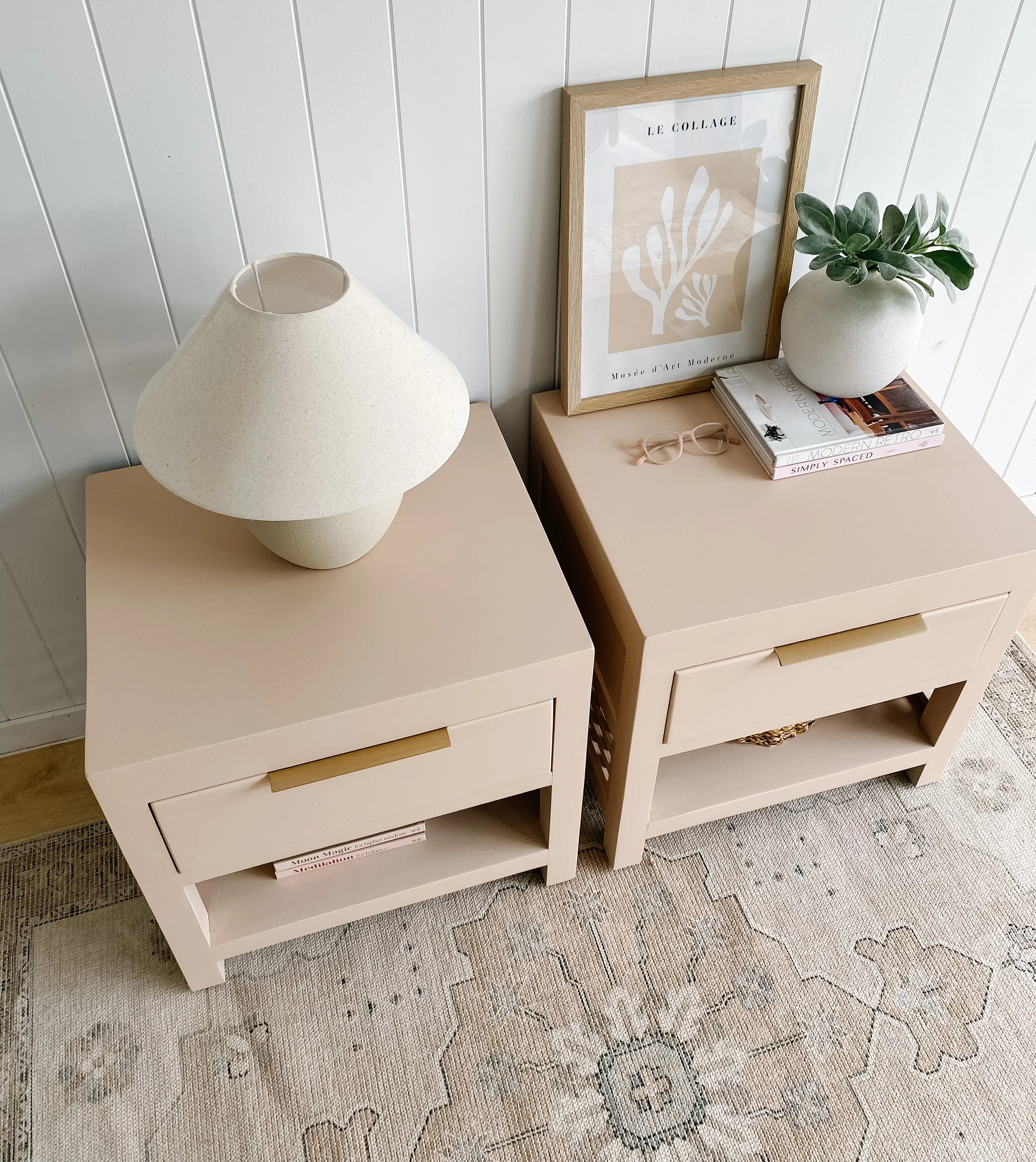 Two beige nightstands with a drawer and lower shelf, placed on a patterned rug next to a white wall panel. The left nightstand has a large, cream-colored table lamp, while the right one holds a potted plant, a framed art print, a pair of glasses, and a stack of books.