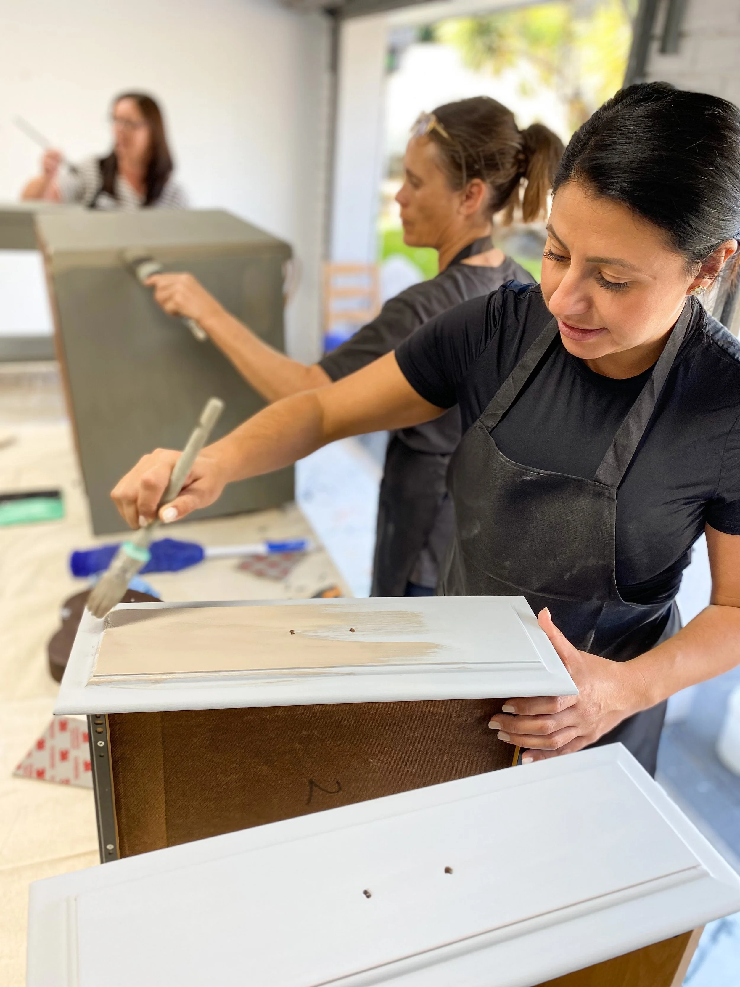 Women painting furniture outdoors, with one woman holding a paintbrush and others working on different furniture pieces.
