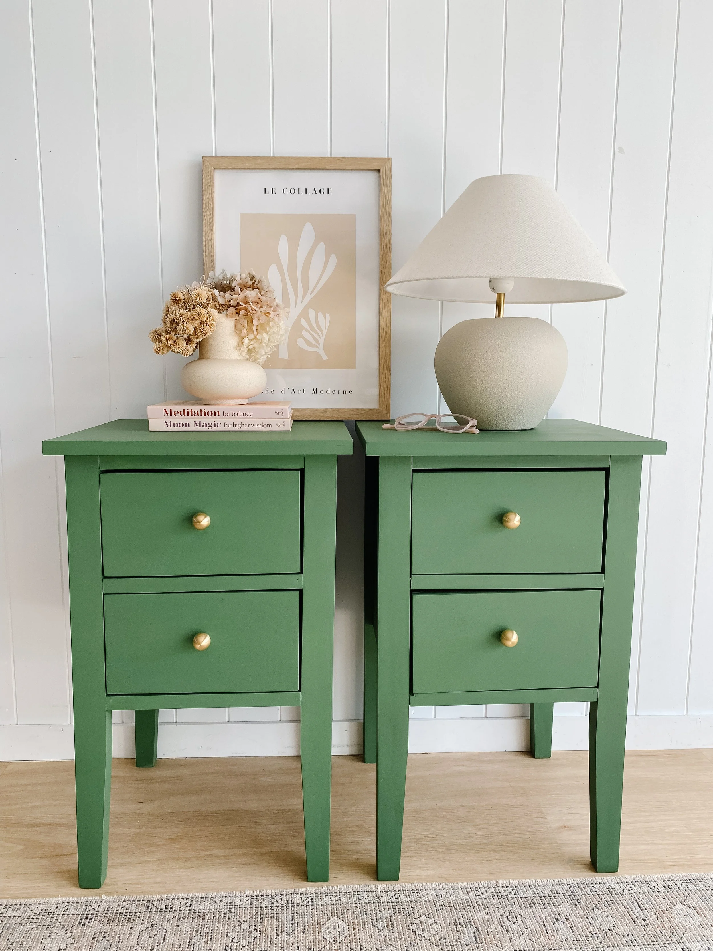 Two green nightstands with two drawers each, decorated with a framed abstract art print, a cream-colored vase with dried flowers, a white table lamp, a pair of glasses, and two books, all set against a white paneled wall and light wood floor.