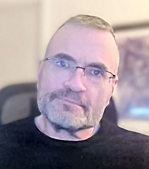 A middle-aged man with glasses, short gray hair, and a beard, wearing a black shirt, sitting in what appears to be a home office or living room.