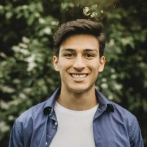 A man with curly dark hair, glasses, and a beard, wearing a dark sweater, sitting indoors with a plant in the background.