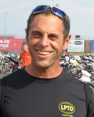 A man with dark wavy hair, wearing clear glasses and a nose ring, seated indoors with a neutral expression, a white wall, a lamp, and a plant in the background.