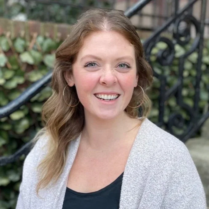 A woman with shoulder-length black hair, smiling, standing among green plants in a bright, indoor setting.