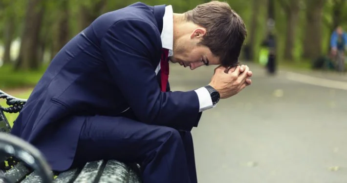 A man in a suit sitting on a park bench, head bowed with hands clasped together, appearing distressed or upset. The background shows trees and a paved walkway.