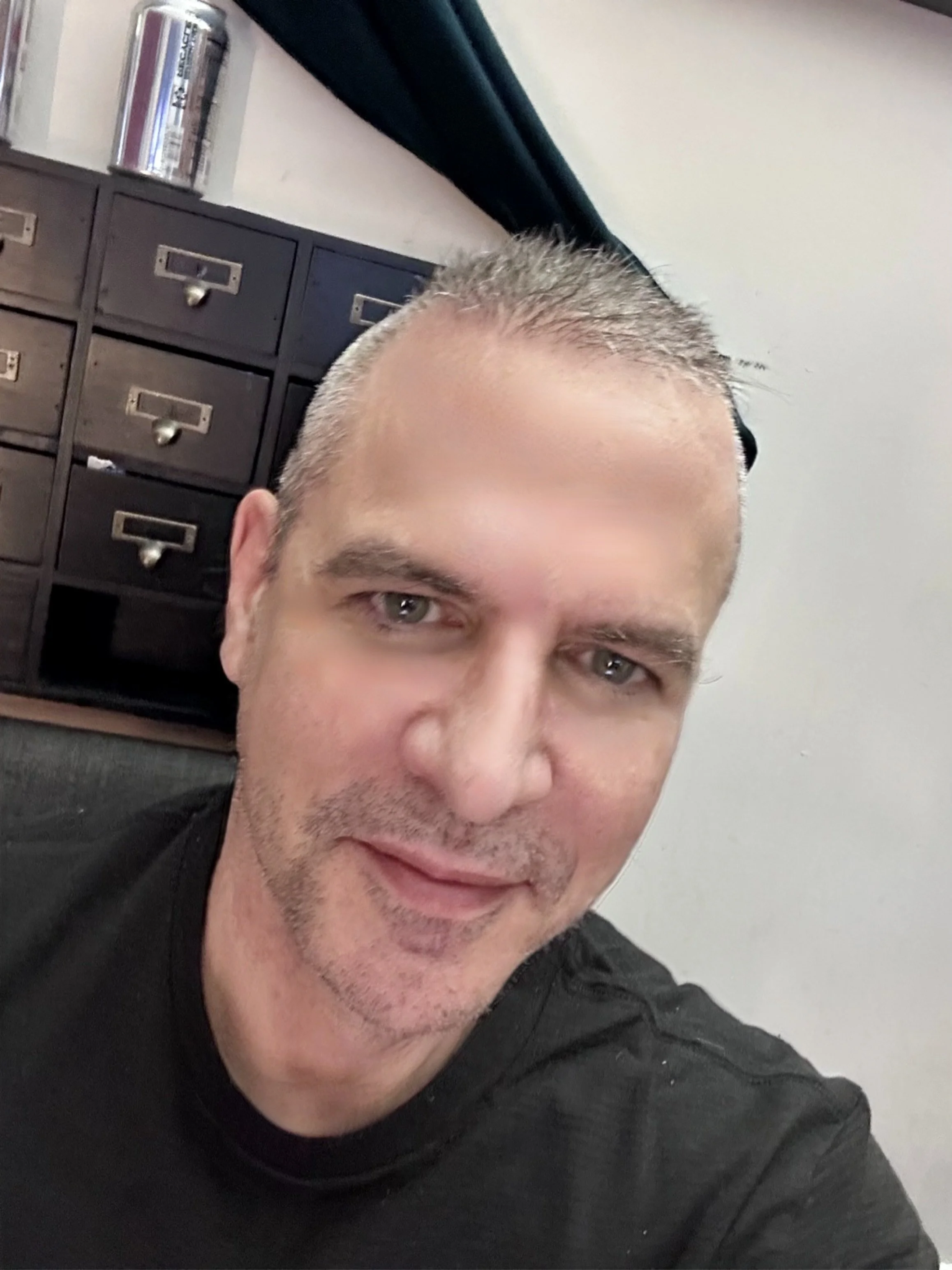 A man's picture, showing his face with short gray hair and a light beard, wearing a black shirt, in an office setting with a desk and small apothecary drawers in the background.