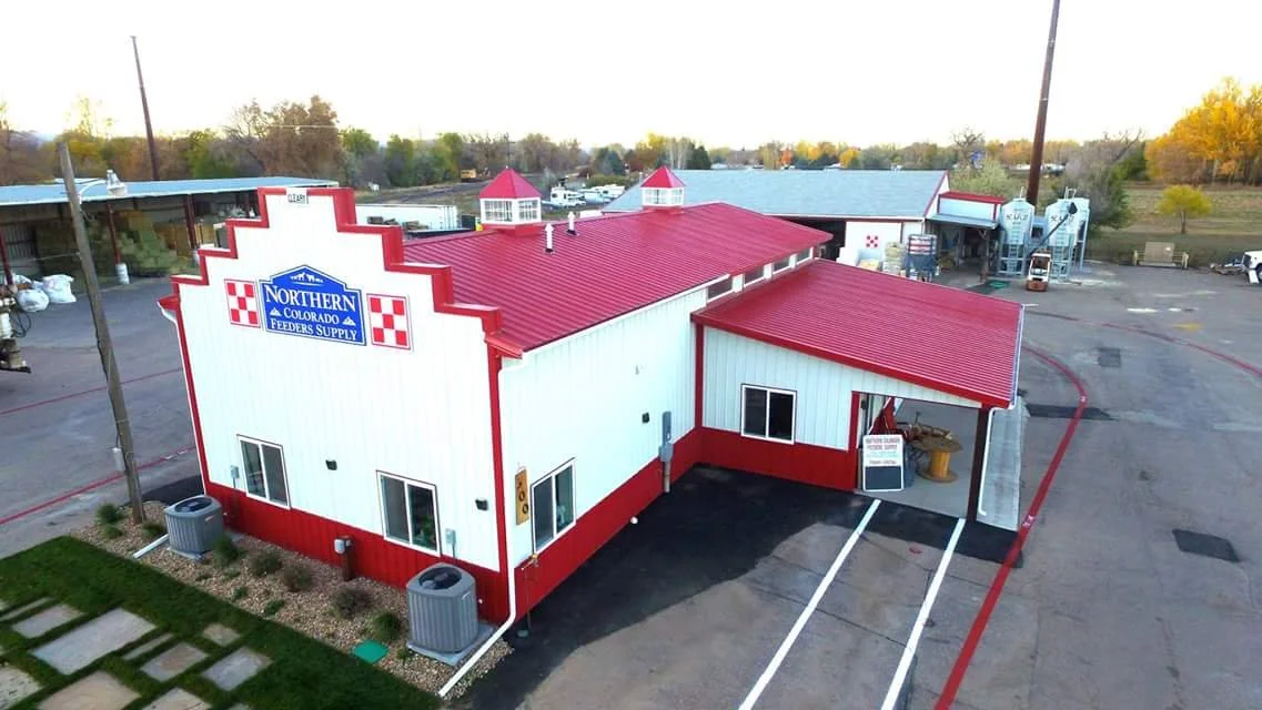 A white building with red trim and a red roof, labeled 'Northern Colorado Feeders Supply,' located in a parking lot with utility poles, surrounding trees, and a rural landscape in the background.