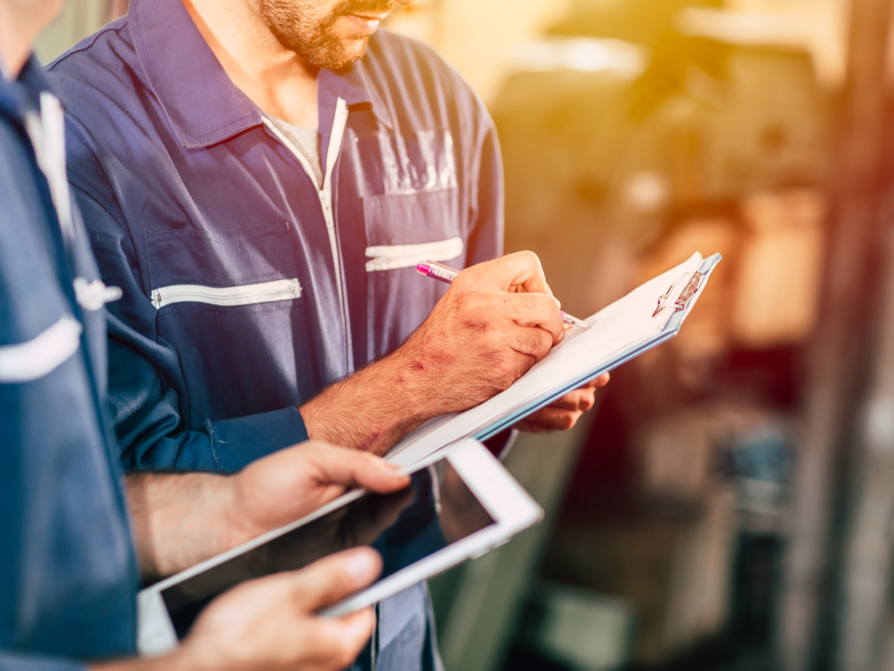 Men in work uniforms taking notes and looking at a clipboard and a tablet.