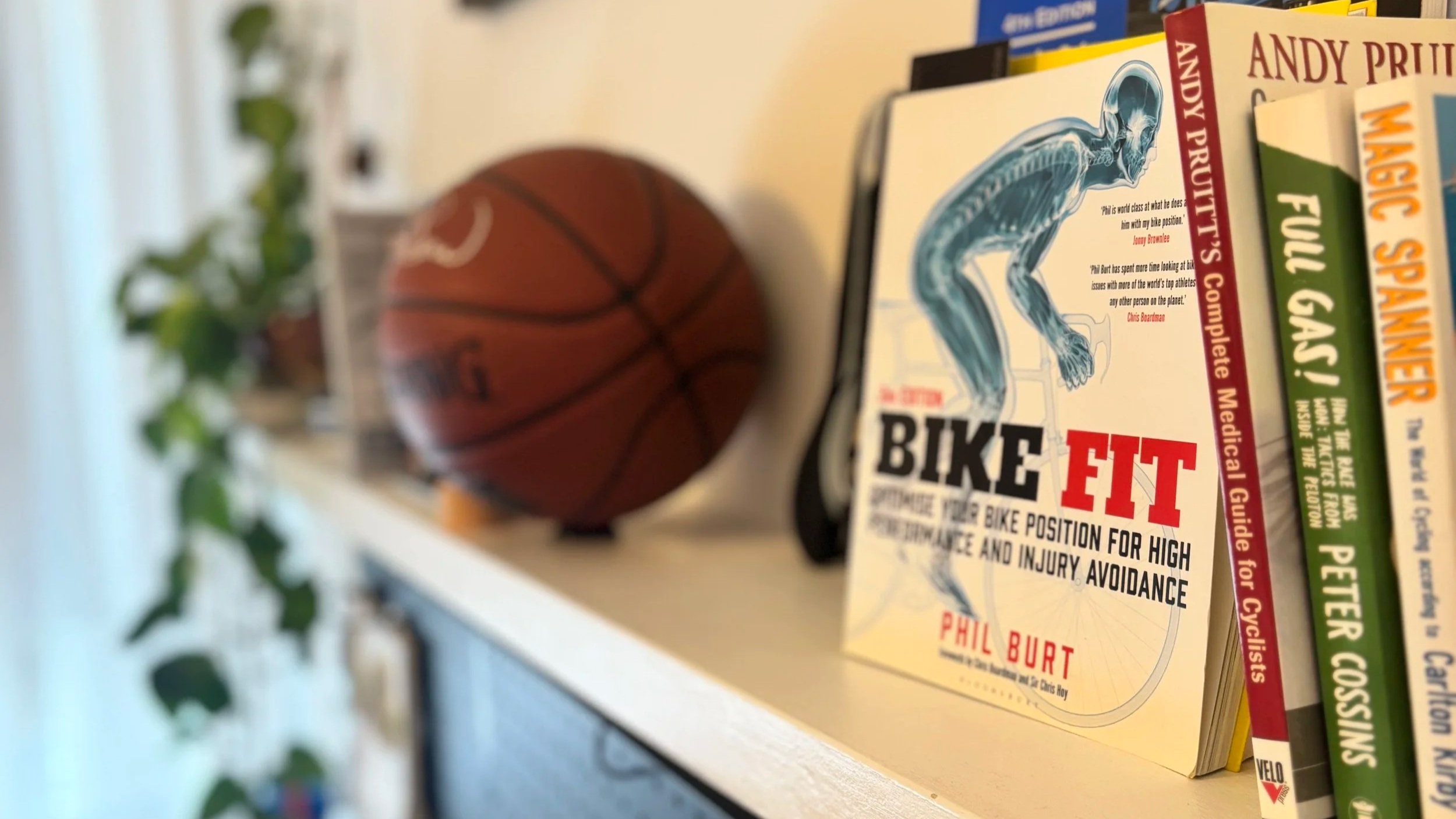 A basketball in a jar in front of a collection of books on a white shelf. The books include titles related to cycling, health, and fitness, with some visible titles like "Bike Fit" and "Andy Pratt".
