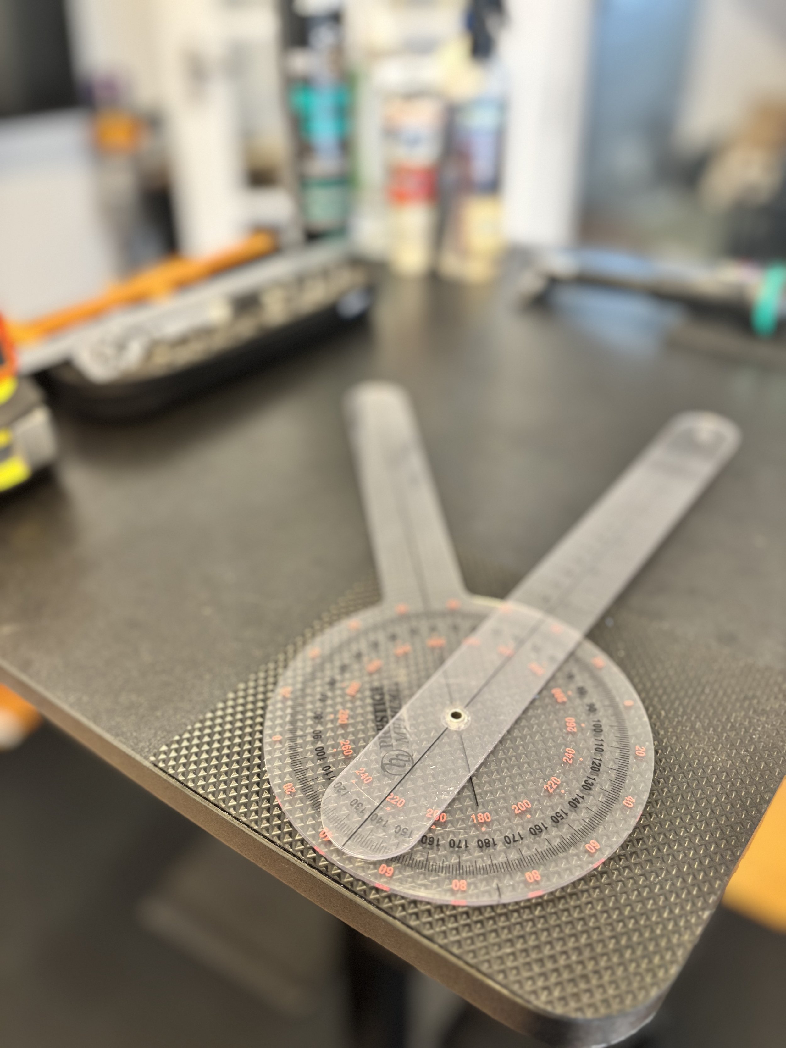 A measuring protractor on a textured metal work surface, with a blurred background of tools and equipment.