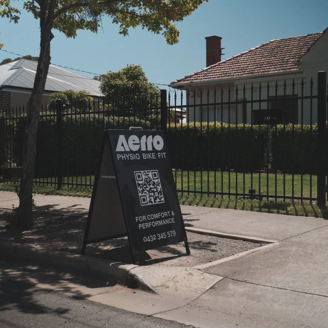 A black A-frame signboard on a sidewalk advertising Aero Physio Bike Fit, with a QR code and contact number, placed next to a tree on a sunny day outside a fenced house.