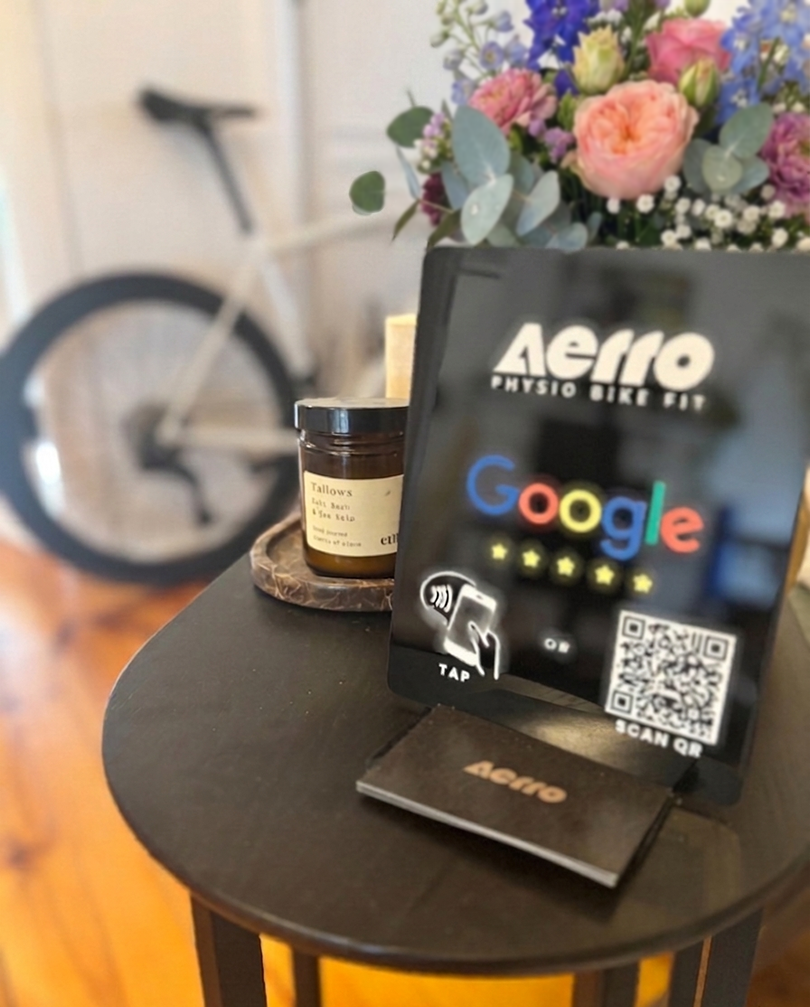 Table with a Google review QR code stand, a small book, a jar labeled "Tallows," and a flower arrangement, with a blurred bicycle in the background.