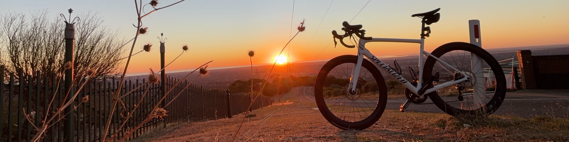A landscape scene at sunset with a white bicycle parked on a dirt path next to a black fence, with dry plants in the foreground and a wide view of the horizon in the background.