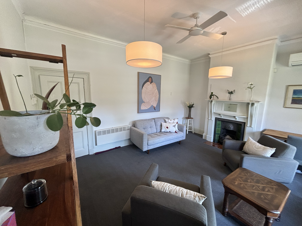 Living room with light-colored walls, ceiling fan, two pendant lights, a gray sofa, a gray armchair, a wooden coffee table, a fireplace with a white mantel, and decorative plants and artwork in Carlton- Drummond Street.