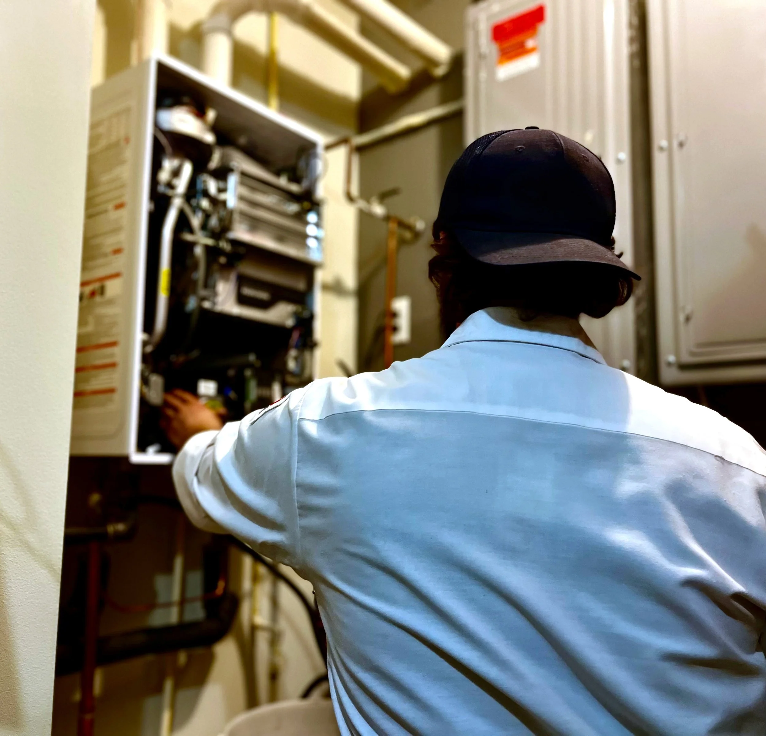 Person wearing a black cap and a light blue shirt, standing in front of an electrical panel with visible wiring and circuit breakers.