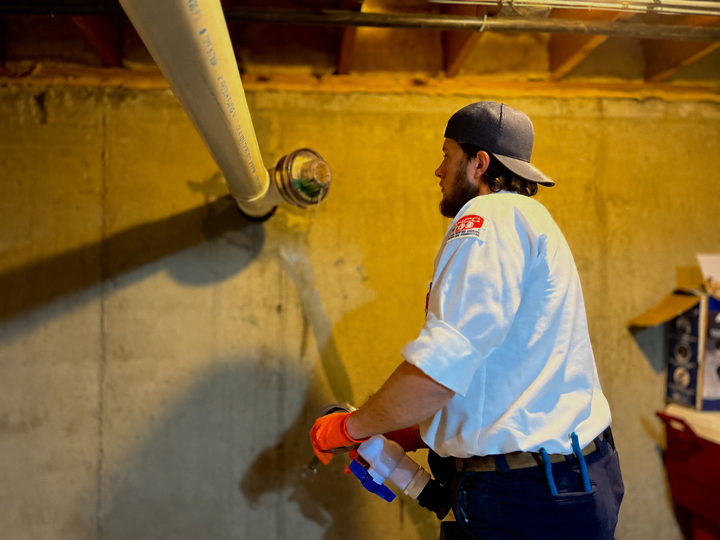 A man wearing a white uniform and a black cap appears to be working on a plumbing project. He is standing next to a concrete wall with a pipe attached to it. He has orange gloves on his hands and is holding a spray bottle. The ceiling above him shows wooden beams.