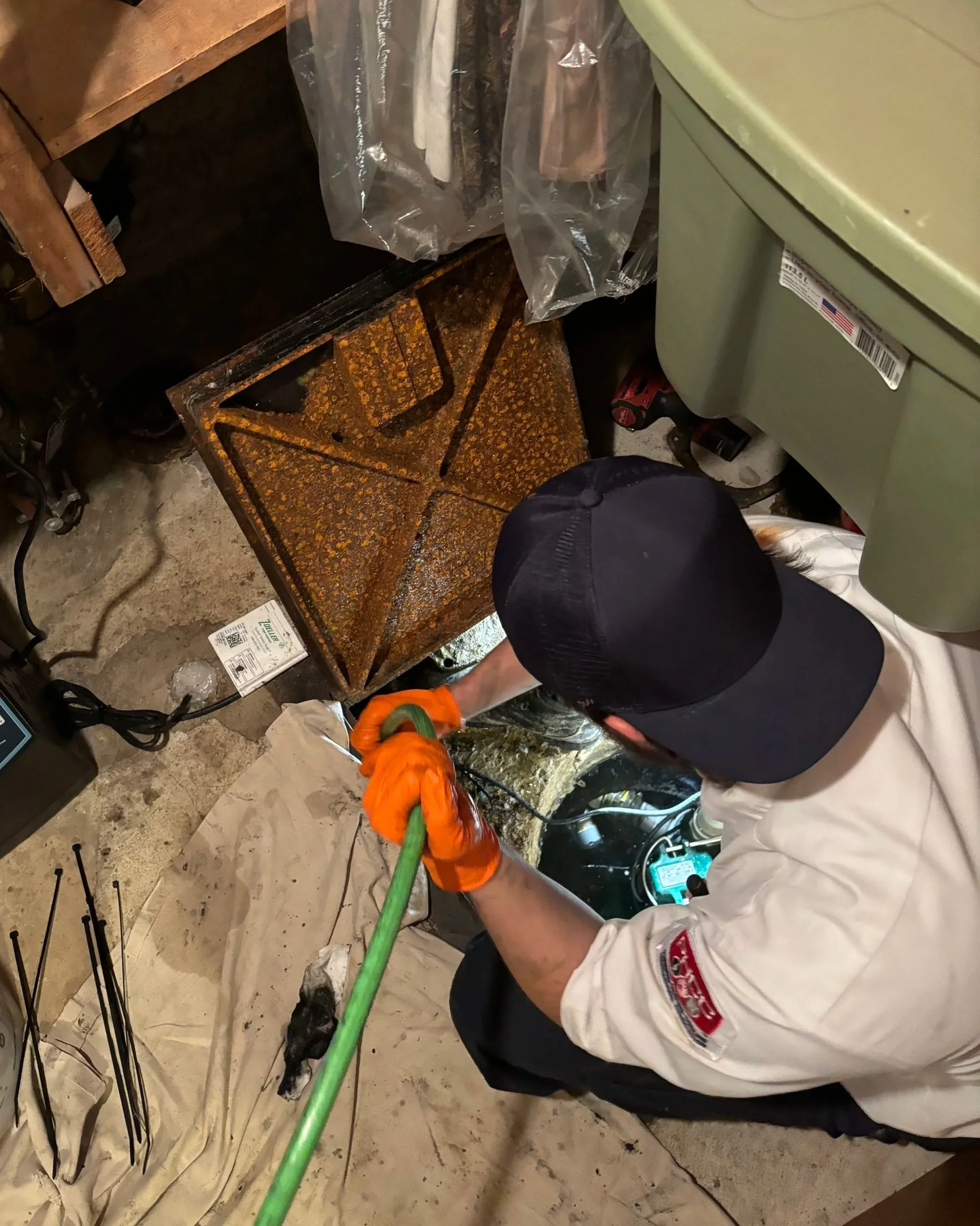 A person working in a basement or utility room, wearing protective gloves and a white uniform, handling a green hose or pipe. They are kneeling or crouching on a concrete floor next to some electrical cords, tools, and a humidity meter or electronic device. There is a rusty metal rack, a plastic bag with hanging items, and a green appliance or generator nearby.