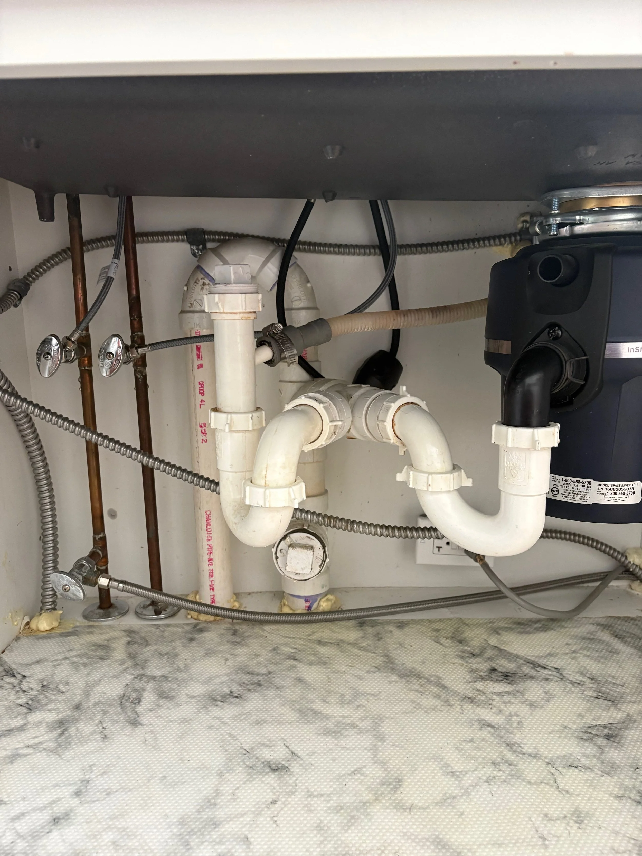 View of plumbing under a kitchen sink, with pipes, valves, and a garbage disposal unit, against a marble countertop