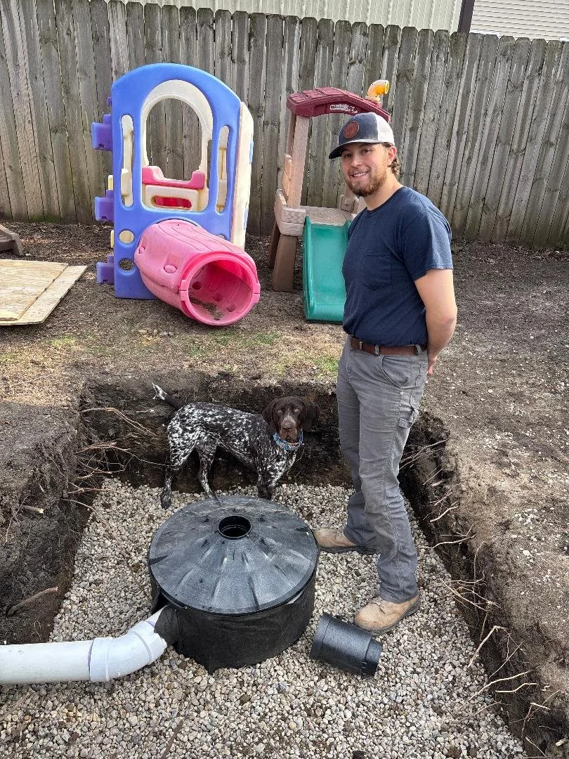 A man standing in a dug hole in the backyard, with a German shorthaired pointer dog inside the hole. There are children's play structures, including a slide and a tunnel, in the background. The man is smiling and looking at the camera, wearing a baseball cap, navy t-shirt, and gray pants.