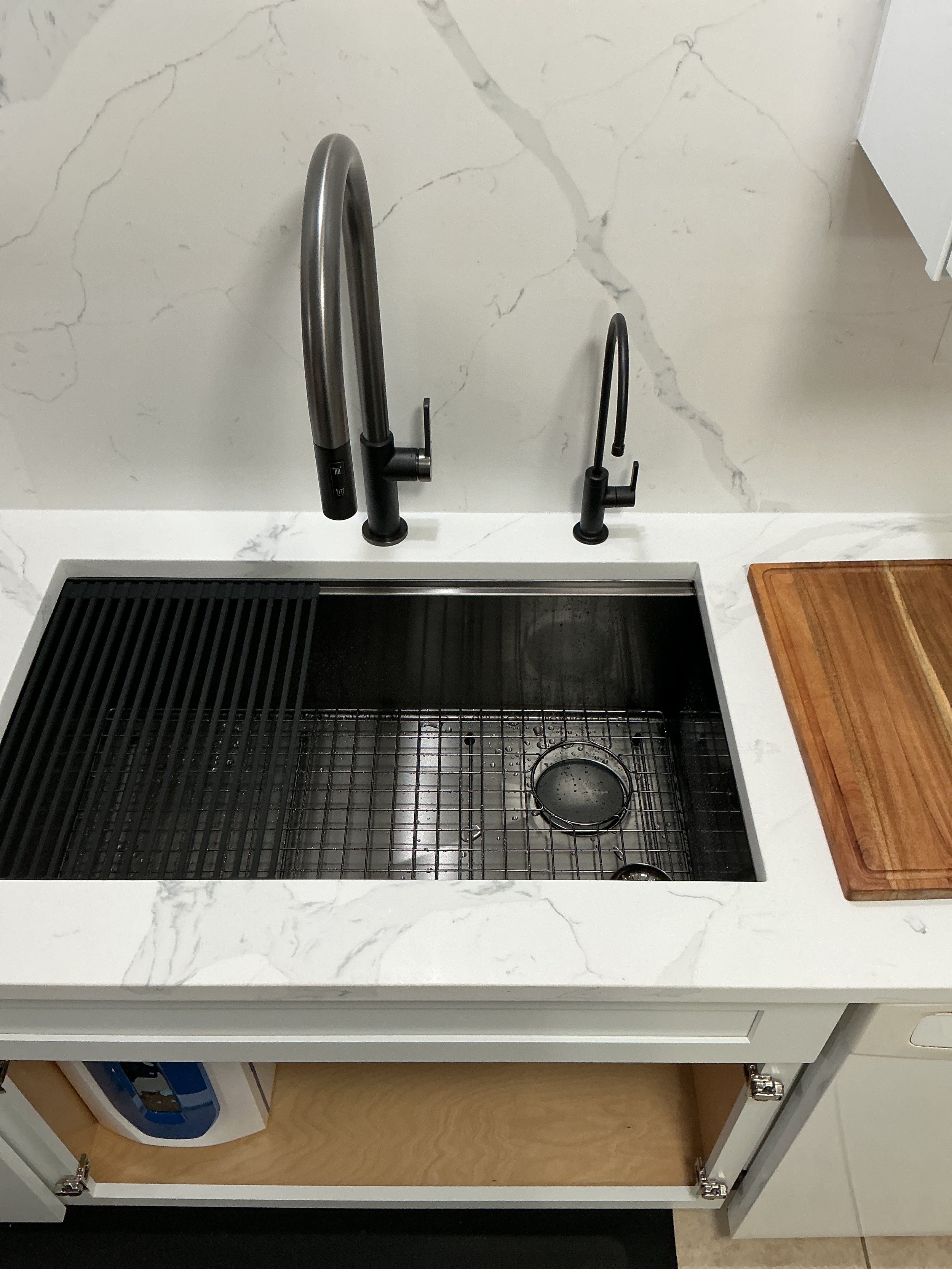 A kitchen sink with a black faucet, a smaller black faucet, a sink with a black grate and utensil holder, a wooden cutting board, and a laundry area visible through an open cabinet door with a washing machine inside.