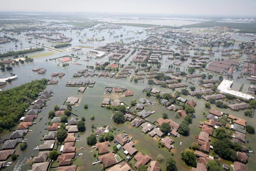 Aerial view of a flooded suburban neighborhood with submerged houses, roads, and trees, showing widespread flooding.