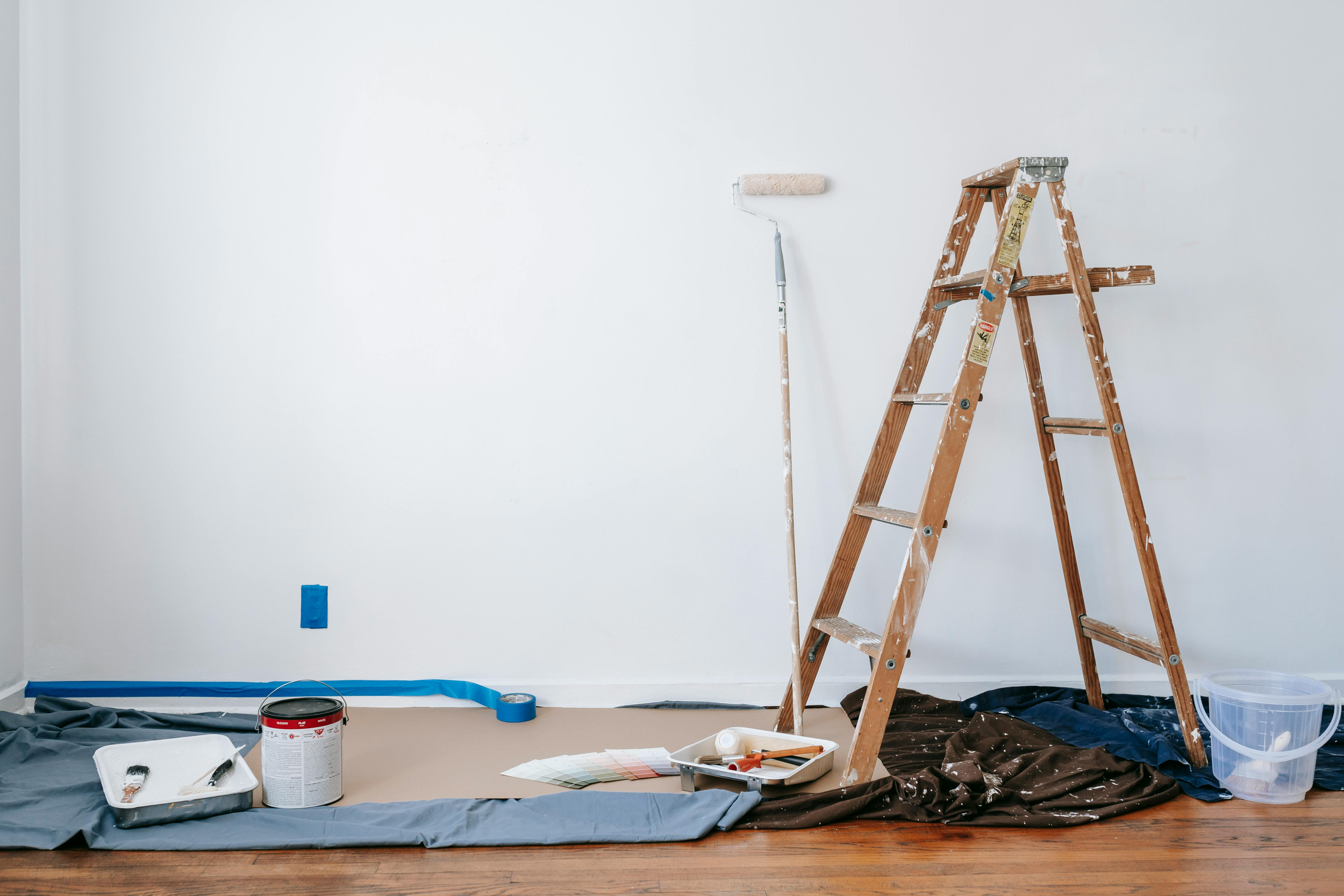 Room being painted with painting supplies, including a paint roller on a roller handle, a ladder, painter's tape, a paint can, paint swatches, and a paint tray, with a drop cloth spread on the floor.